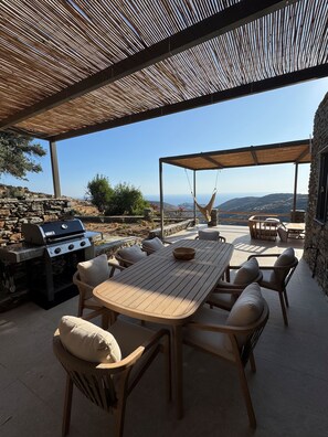 An outdoor patio with a wooden table and chairs, a grill, and a swing chair. - Mythic Villa (Sifnos)