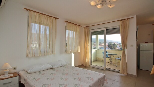 A bedroom with a bed, a nightstand, a window with curtains, a balcony with a table and chairs, and a refrigerator. - Charming studio Ljubica (Punat)