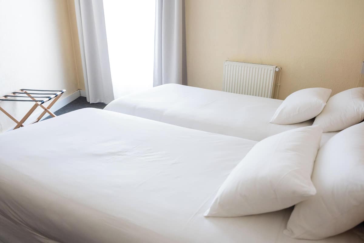 Una camera d'albergo con due letti, lenzuola bianche, un termosifone e uno stendibiancheria.