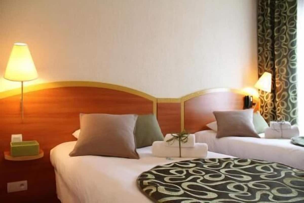 Camera d'albergo con testiera in legno, due letti, lampade da comodino, una coperta verde e nera fantasia e una piccola scatola verde sul comodino.
