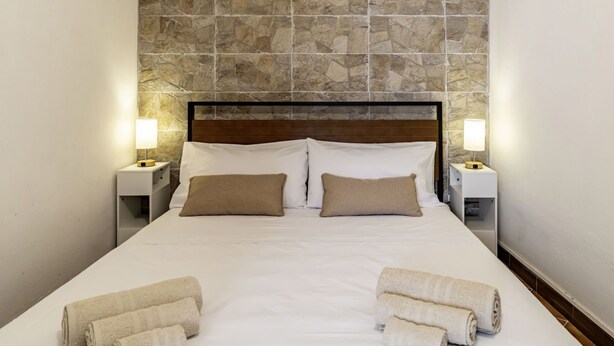 A neatly made bed with white linens and two pillows, flanked by two bedside tables with lamps, set against a stone wall.