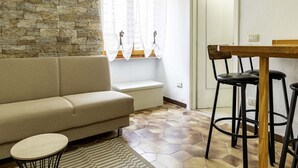 A living room with a stone wall, a sofa, a small table, a window with curtains, and a wooden bar stool.
