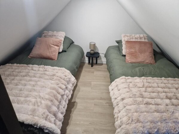 A small, cozy room with two beds, each with a fluffy blanket and a pillow. There is a small table with a lamp between the beds.