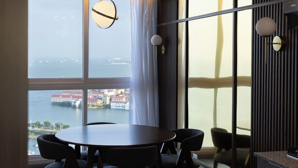 A modern room with a round table and six chairs, large windows with a view of a body of water and buildings, and two wall-mounted light fixtures. - 1 bdr apartment with Ocean View and Social area w/ pool (Avenida Balboa)