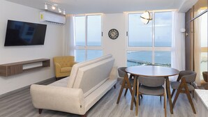 A modern living room with a sofa, a TV mounted on the wall, a dining table with chairs, and large windows offering a view of the sea. - 1 bdr apartment with Ocean View and Social area w/ pool (Avenida Balboa)