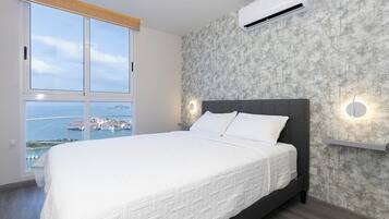 A bedroom with a large bed, an air conditioner, a nightstand, and a view of a body of water through a window.