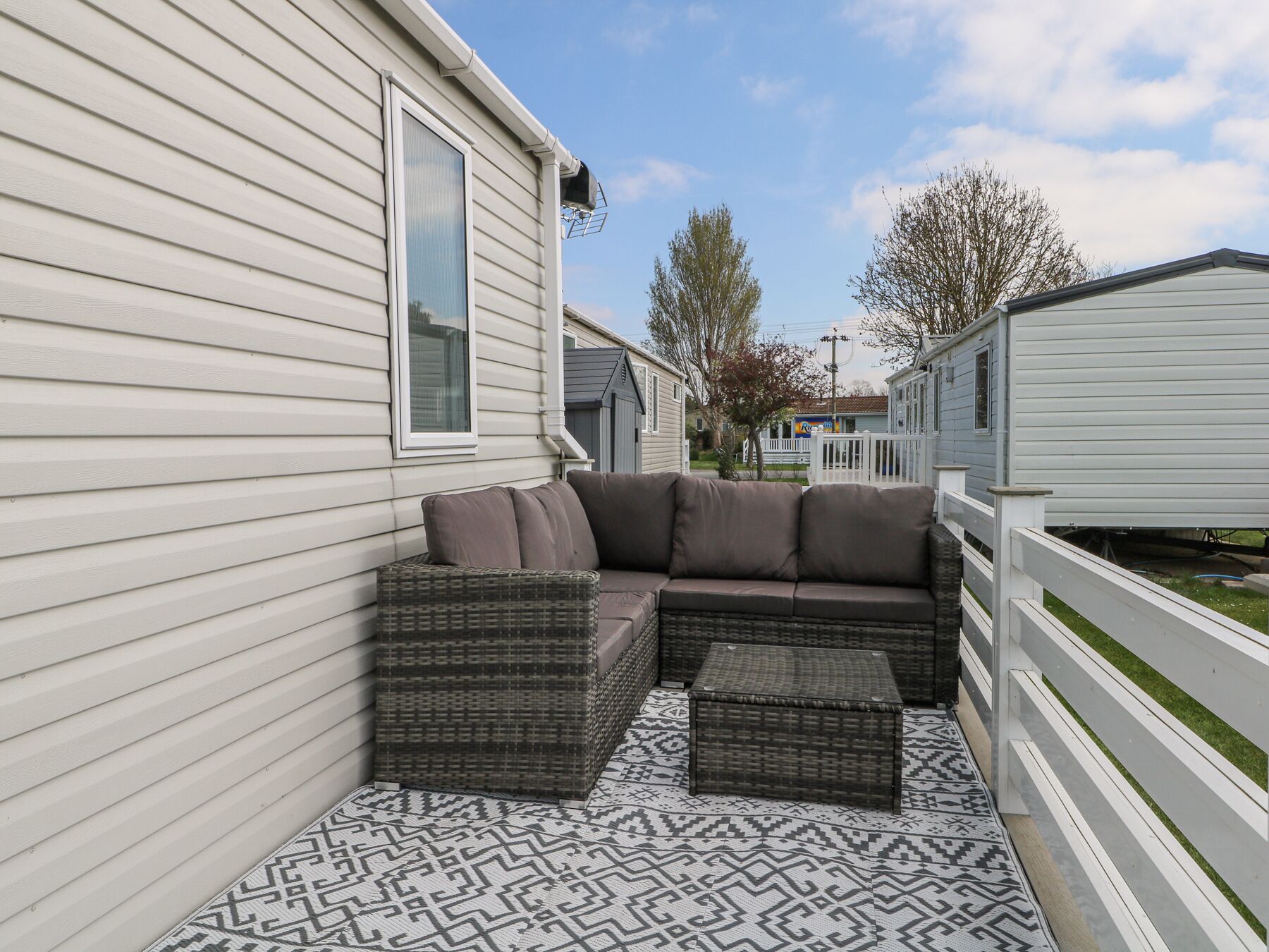 Un patio cubierto con un sofá modular y muebles de mimbre, contiguo a una casa móvil con revestimiento blanco.