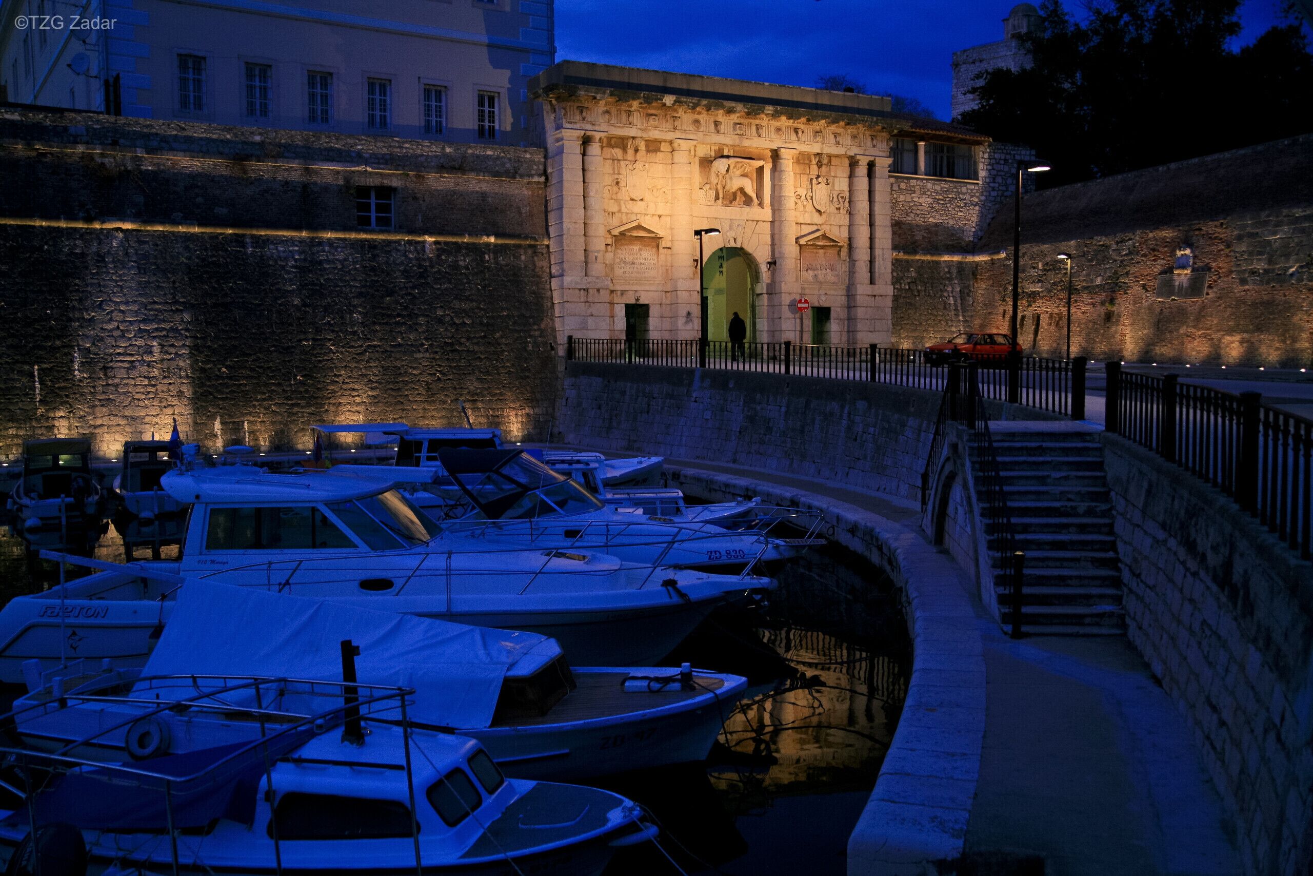 A marina with boats docked, a stone wall, and a building with illuminated windows and a statue.