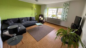 Ein Wohnzimmer mit einem Ecksofa, einem kleinen Couchtisch, einer Lampe, einer Pflanze und einem Fernseher.