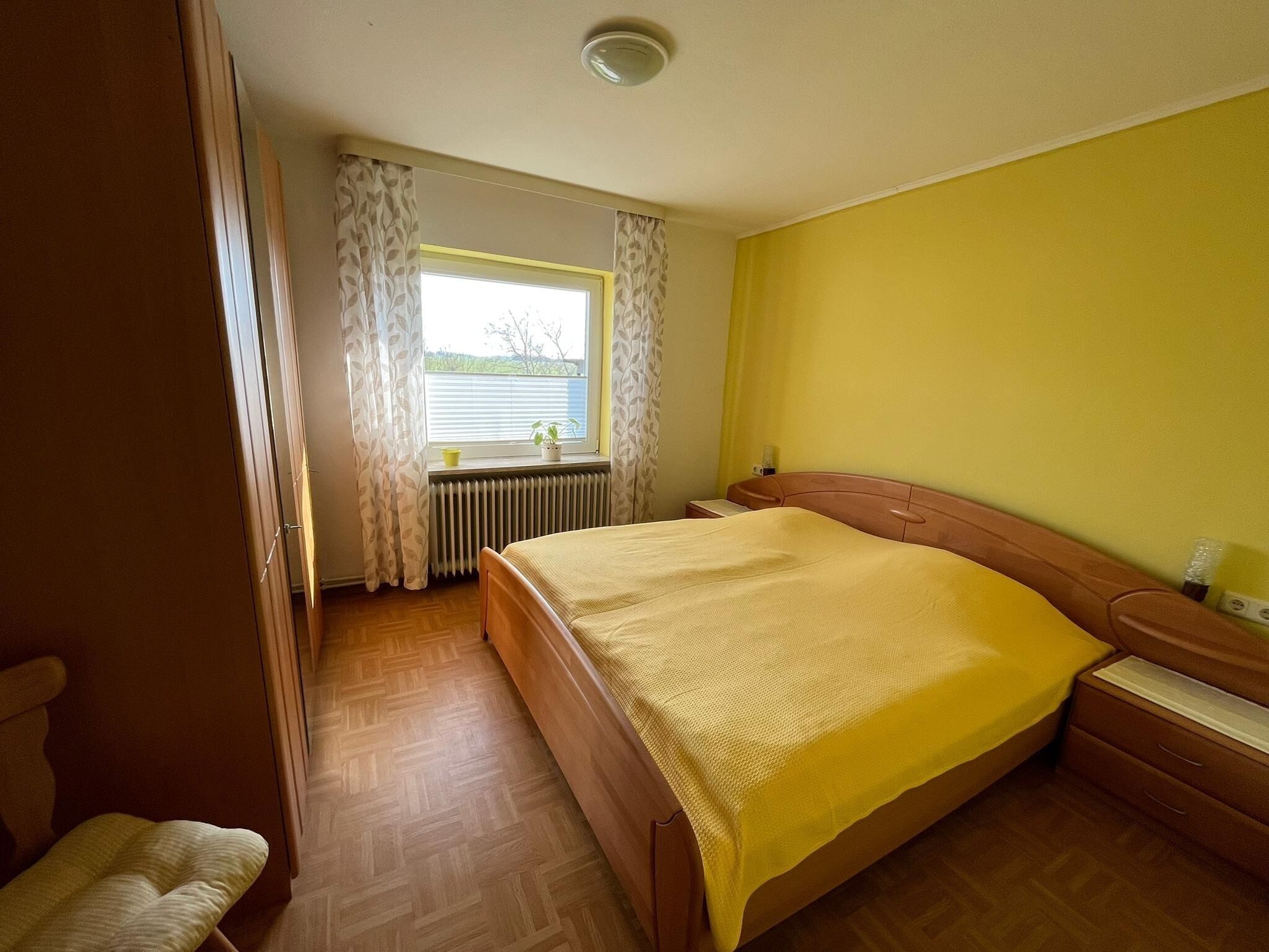 A bedroom with a bed, wooden headboard, a window with curtains, a radiator, and a bedside table.