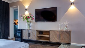A modern living room with a flat-screen TV mounted on the wall, a wooden herringbone-patterned cabinet, a glass coffee table, and a vase with flowers on a side table. - Langschläfer - New: Apartment Langschläfer 4th floor, best location on the beach (Westerland)