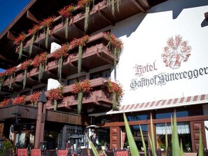 A hotel facade with balconies adorned with red flowers, a prominent sign with a coat of arms, and outdoor seating areas. - Standard double room - Hotel Gasthof Mitteregger (Kaprun)