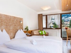 A hotel room with a large bed, a wooden headboard, a view of the city, and a balcony with a table and chairs. - Standard double room - Hotel Gasthof Mitteregger (Kaprun)