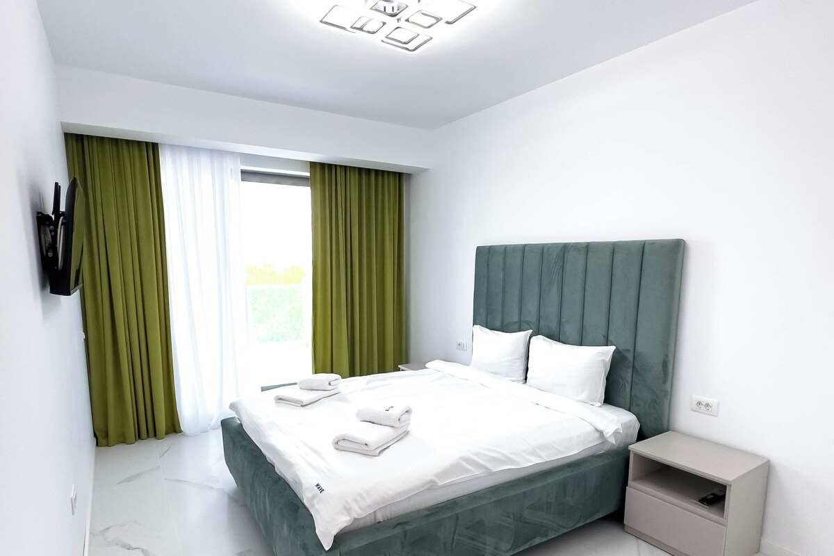 A modern bedroom with a large bed, green curtains, a bedside table, and a nightstand.