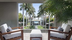 Habitación de hotel moderna con dos sillas de madera, un taburete blanco y vistas a palmeras y una piscina.