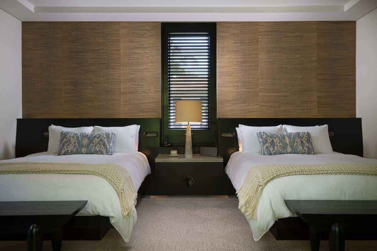 Et hotelværelse med to senge, et natbord, en lampe og et vindue med persienner.