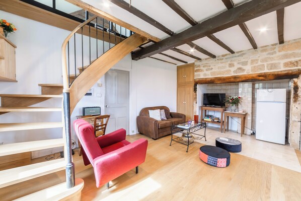 Un vaste espace de vie comprenant un escalier en bois, un fauteuil rouge, un canapé marron, une petite table et un coin cuisine équipé d’une télévision et d’un réfrigérateur.