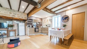 Een ruime keuken met een stenen muur, houten balken en moderne apparatuur. Er is een eethoek met een tafel en een bank, en een gezellig zithoekje met een bank en kussens.