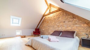 Een slaapkamer met een schuine kap, een stenen muur, een bed met kussens, een rode stoel, een klein tafeltje en een mand op de vloer.