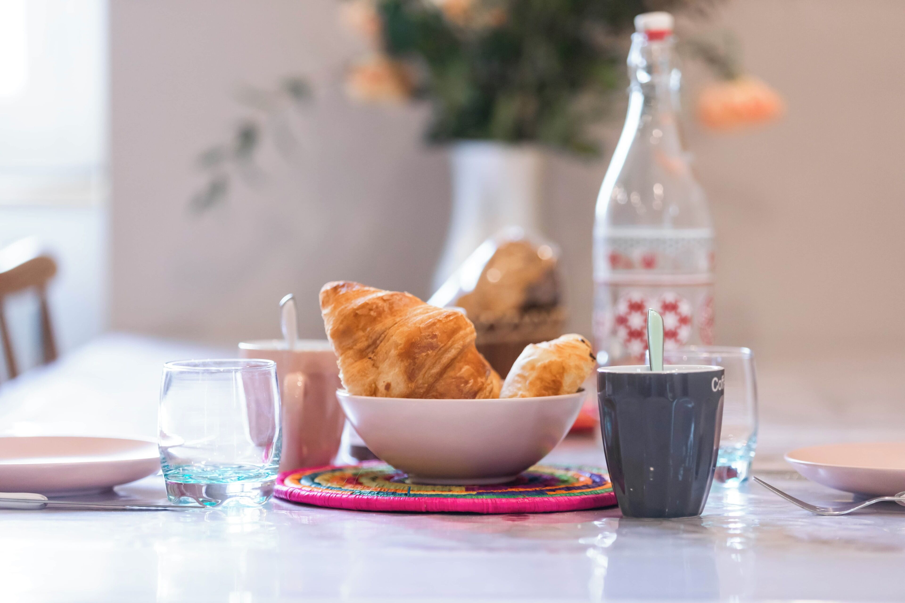 Ein Tisch mit einer Schale Croissants, einem Glas Wasser, einer Tasse und einem Löffel sowie einer Flasche.