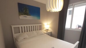 A neatly made bed with white linens, a bedside lamp, and a painting of a beach scene on the wall. - Spacious Family Apartment in Málaga, near historic center (Málaga)