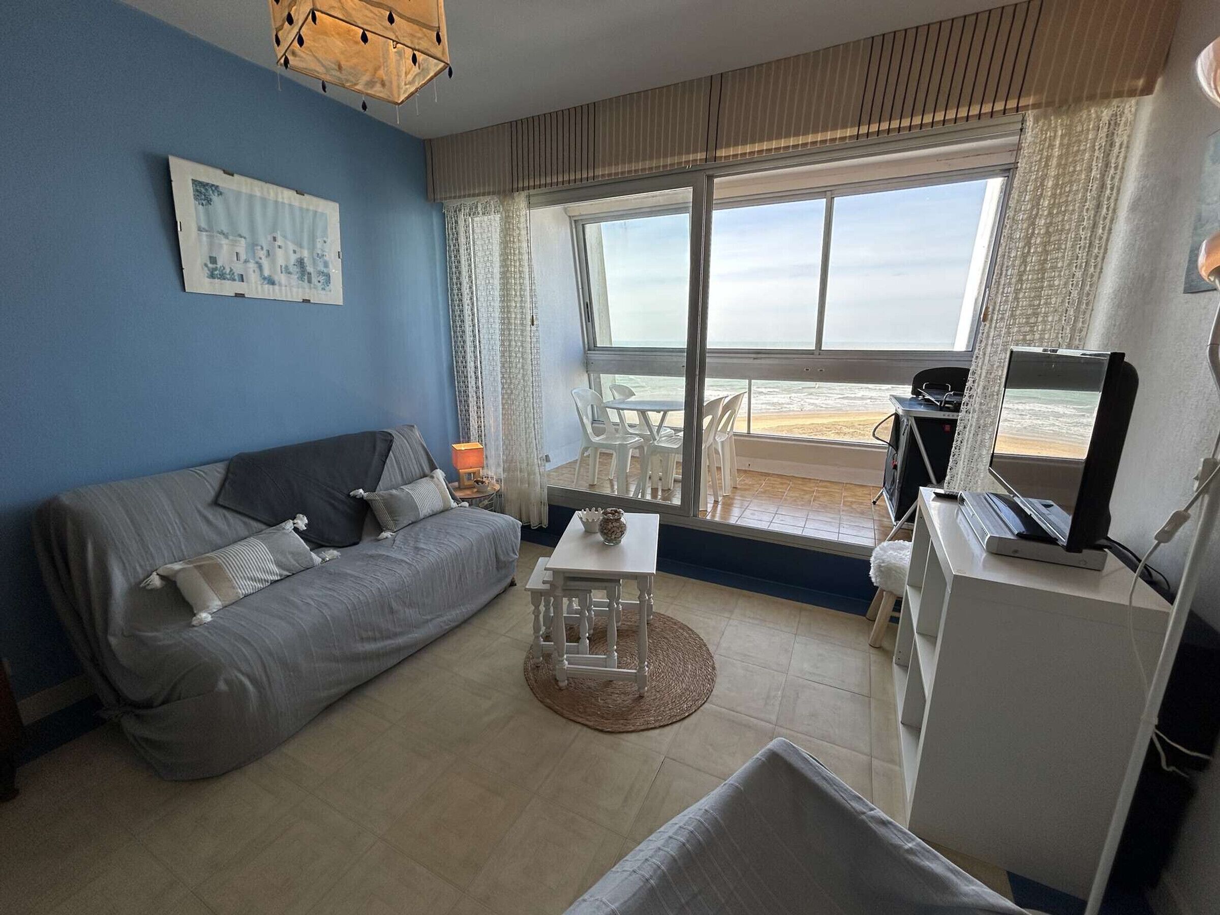 A living room with a sofa, a small table, and a TV on a desk. There is a window with a view of the beach.