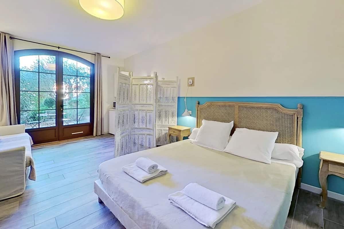A hotel room with a large bed, a desk, a chair, a window with curtains, and a view of greenery outside.