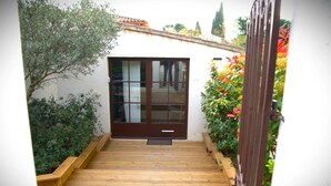 Une terrasse en bois mène à une porte marron dotée de panneaux de verre, le tout entouré de verdure et bordé d’un mur blanc.