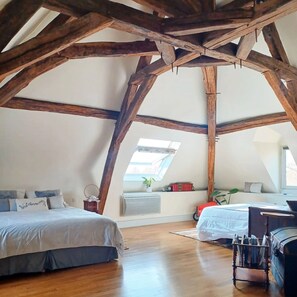 Un dormitorio con techo inclinado, vigas de madera, tragaluz, cama, escritorio y cómoda.