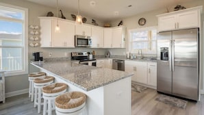 A modern kitchen with white cabinets, a stainless steel refrigerator, and a granite countertop. - Still Waters - Waterfront - NEW Design Build Home - Ocean View (North Topsail Beach)