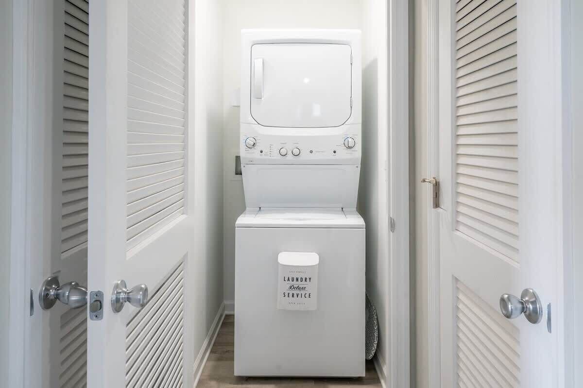 A laundry room with a front-loading washing machine and dryer, a door with a handle, and a sign indicating laundry service.