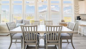 A dining area with a large table and chairs, a chandelier, and a view through large windows. - Still Waters - Waterfront - NEW Design Build Home - Ocean View (North Topsail Beach)