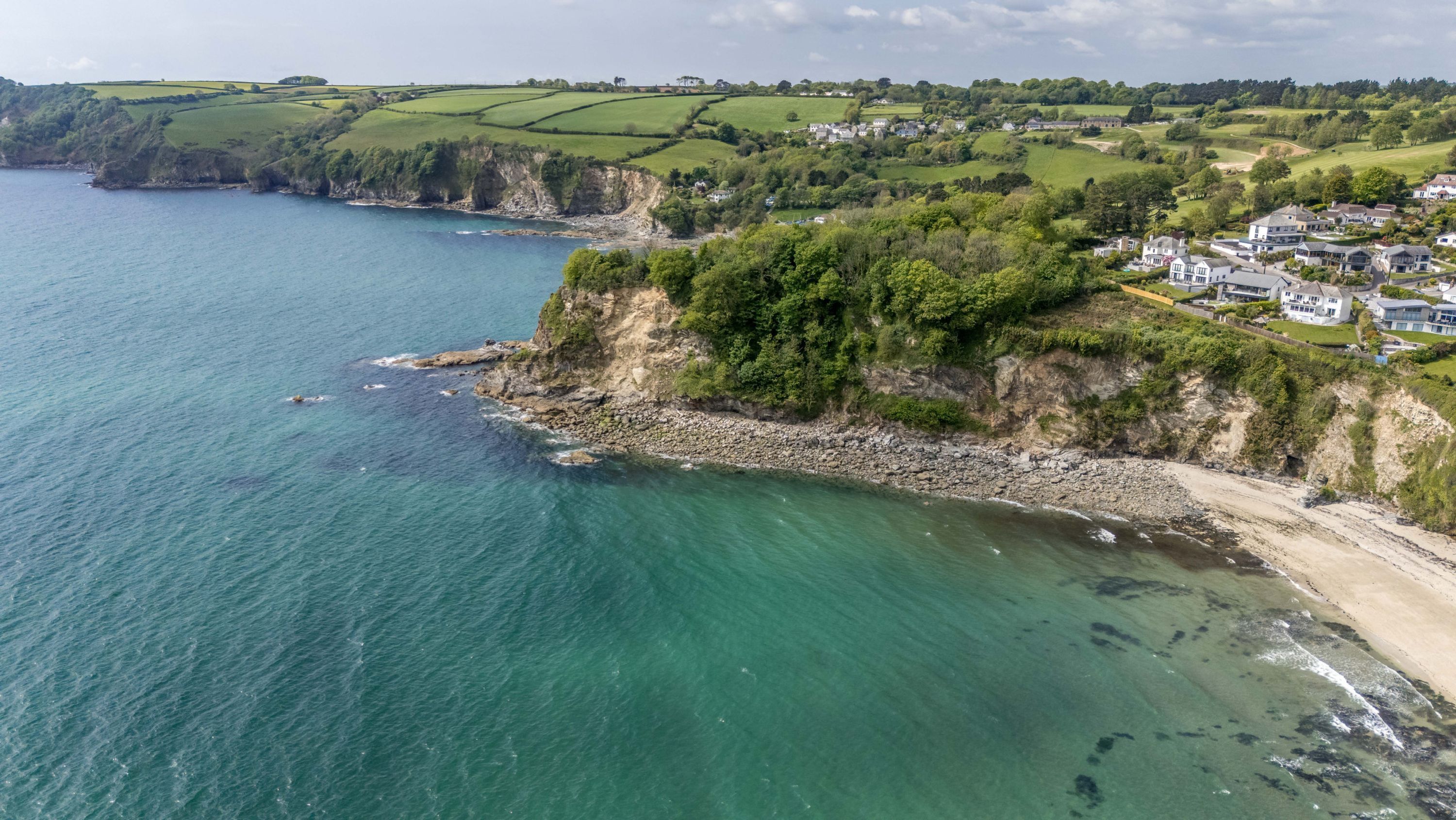 A coastal town with a rocky shoreline, green fields, and houses with visible driveways.