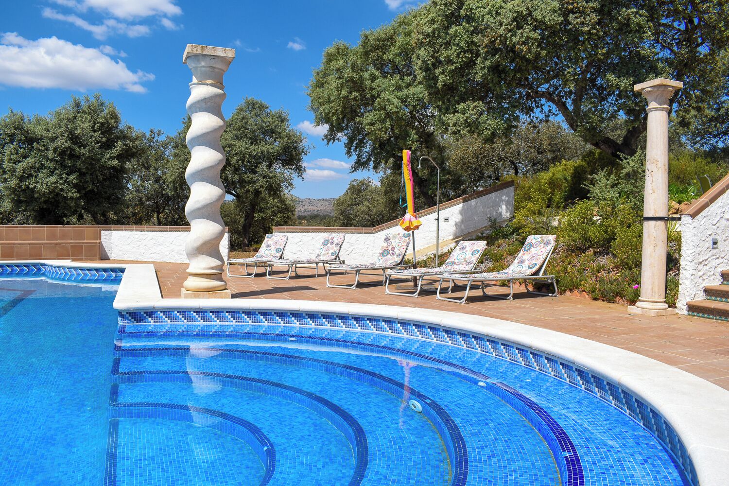 A swimming pool with a wavy edge, surrounded by lounge chairs and a stone column.