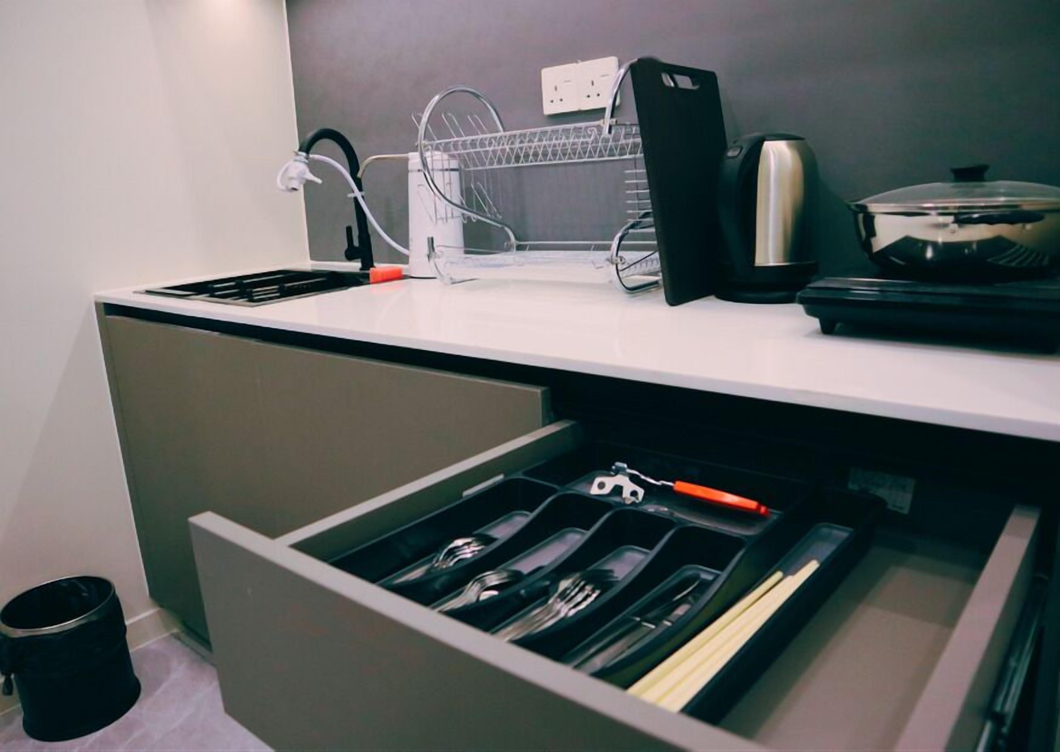 A kitchen with a white countertop, a dish rack, a kettle, and a drawer with utensils.