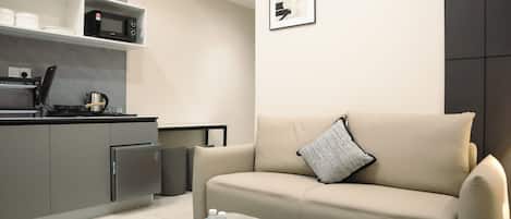 Un soggiorno moderno con un divano beige, un tavolino con bottiglie d'acqua e un telecomando, angolo cottura con forno a microonde e una cornice con un'immagine appesa al muro.