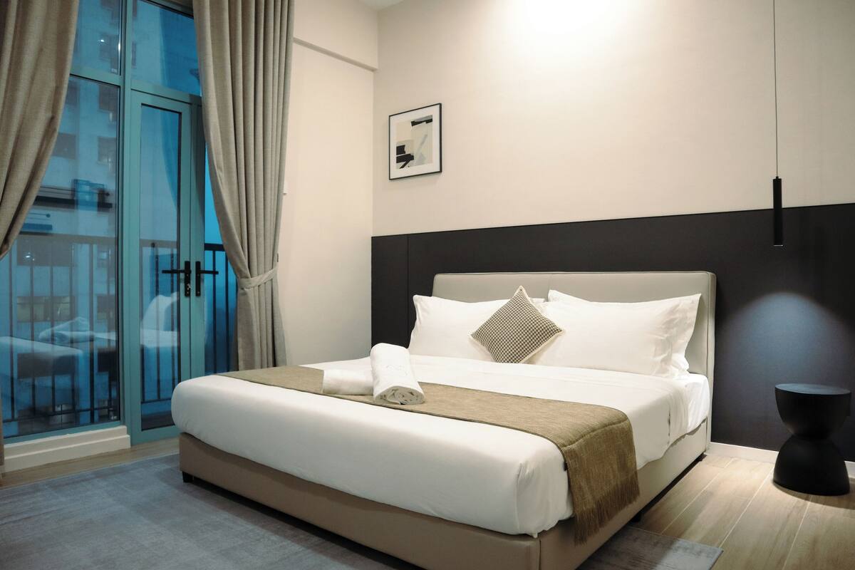 A modern hotel room with a large bed, a view through a glass door, and a small side table.