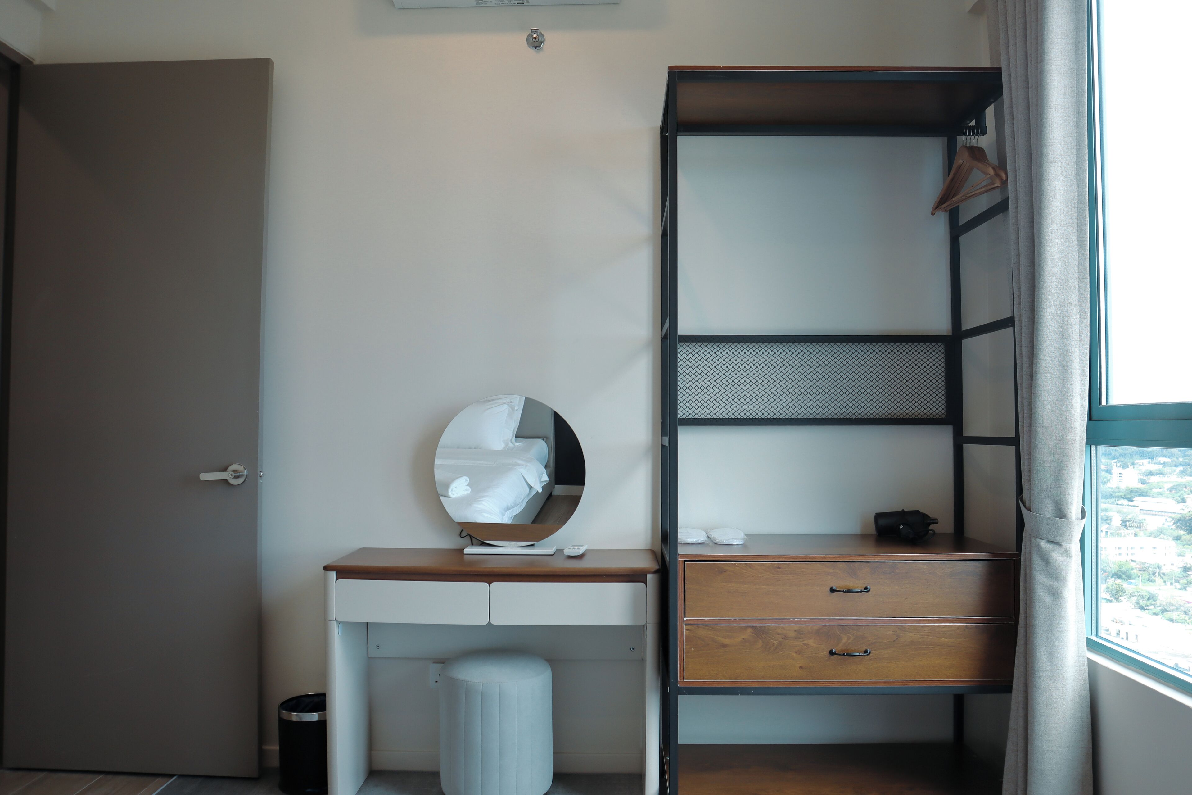 A compact hotel room with a wooden desk, a mirror, a small wardrobe, and a window with a view.