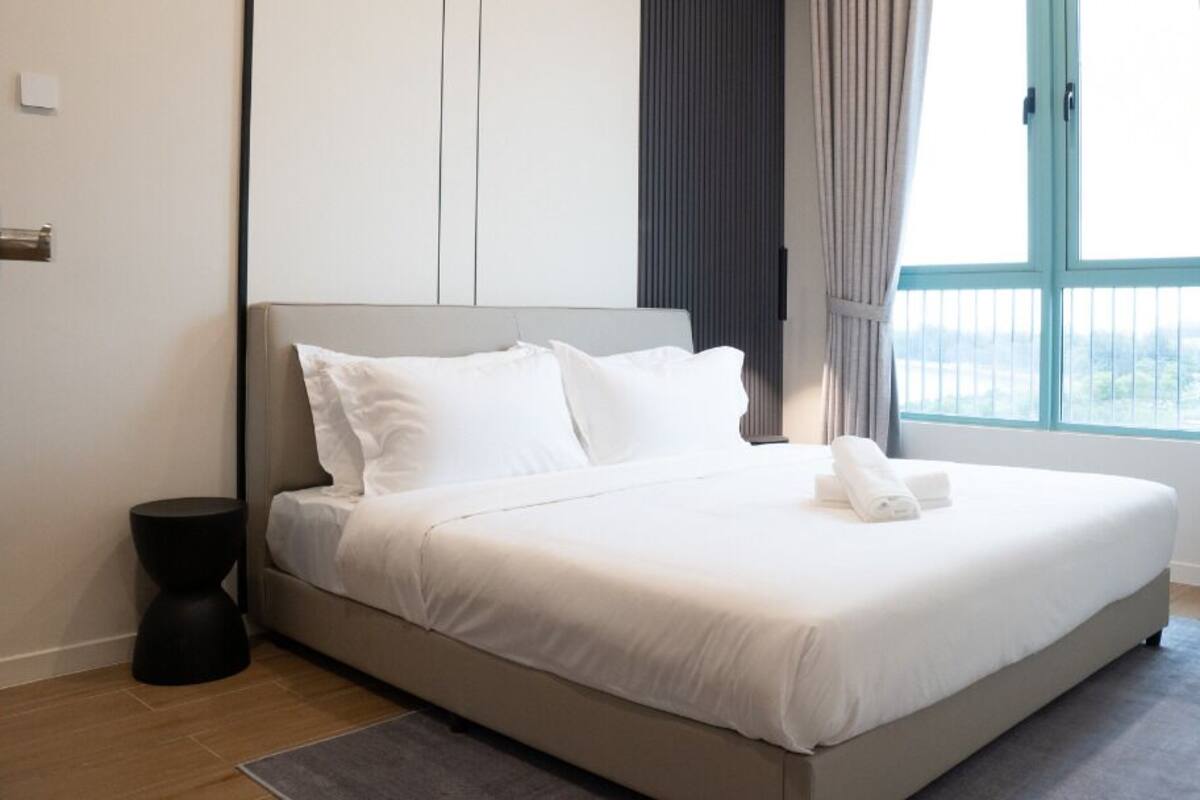A modern hotel room with a large bed, a small side table, and a view of the outdoors through a large window.