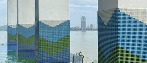 A swimming pool with a tiled design, large concrete pillars, and a view of a distant building across the water.