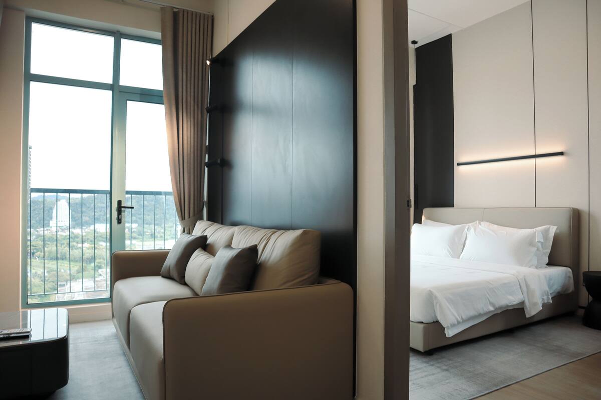 A modern hotel room with a large window, a sofa, a bed, and a nightstand.