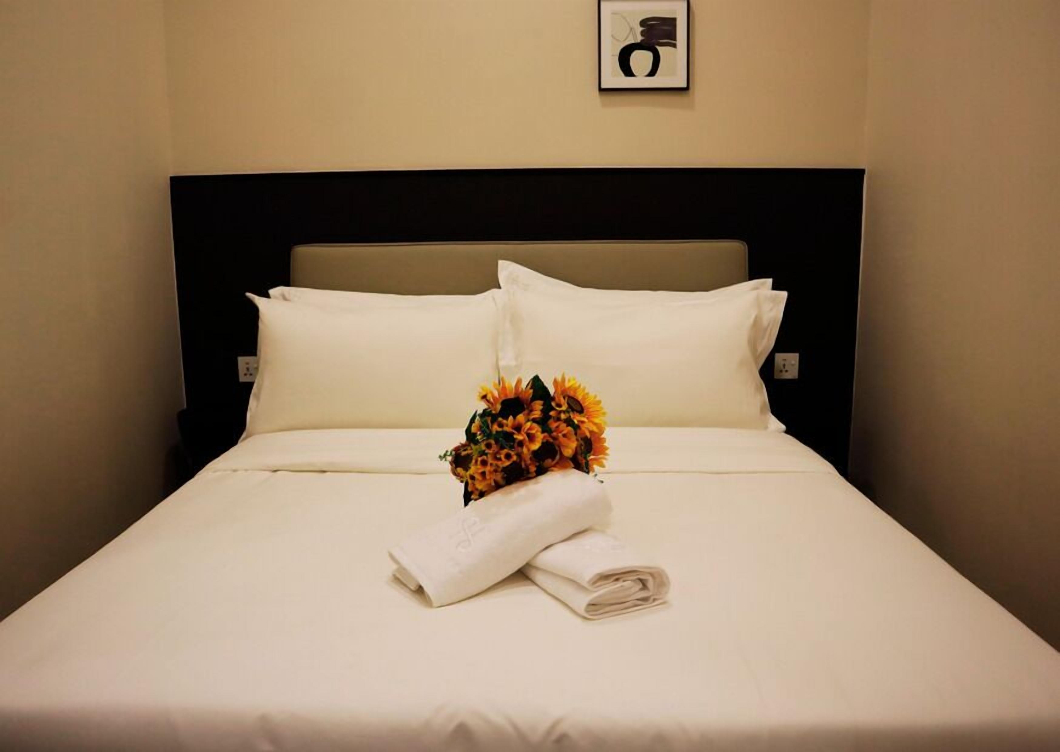 A neatly made bed with white linens, a folded towel, and a bouquet of sunflowers.