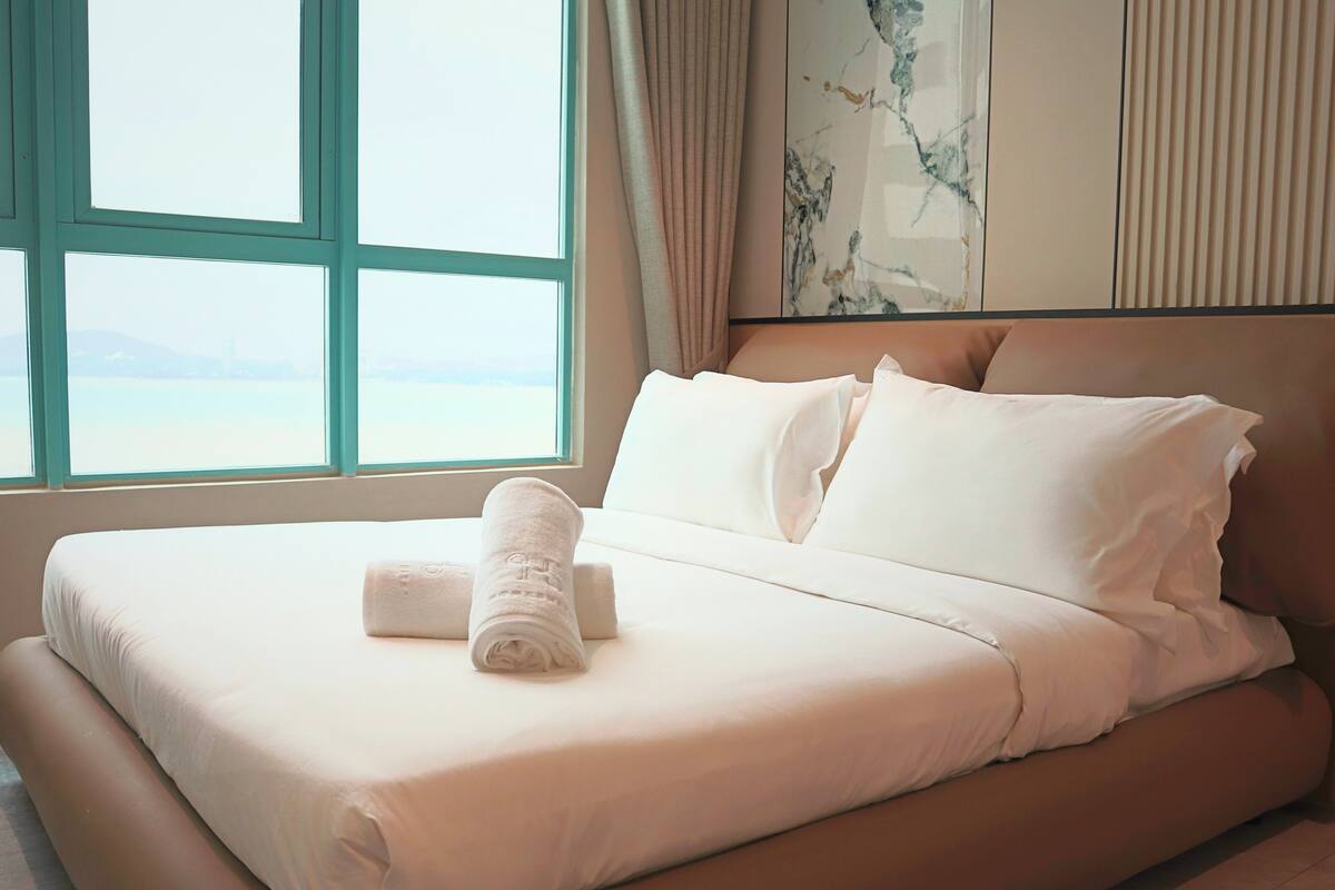 A hotel room with a large bed, white bedding, and a view of the sea through a window.