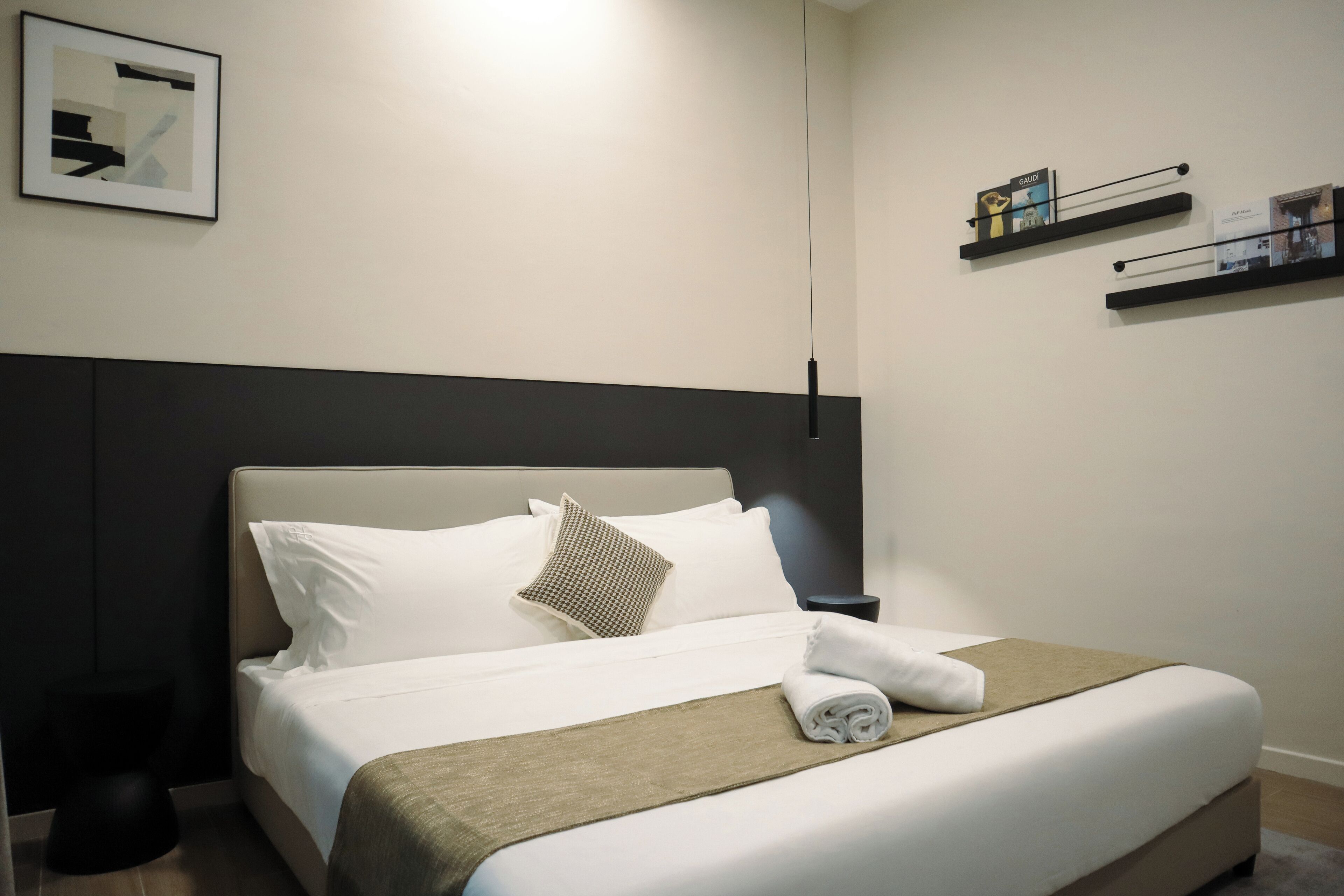 A neatly made bed with a white and brown bedspread, a rolled towel, and a pillow. There is a black nightstand with a lamp beside the bed.