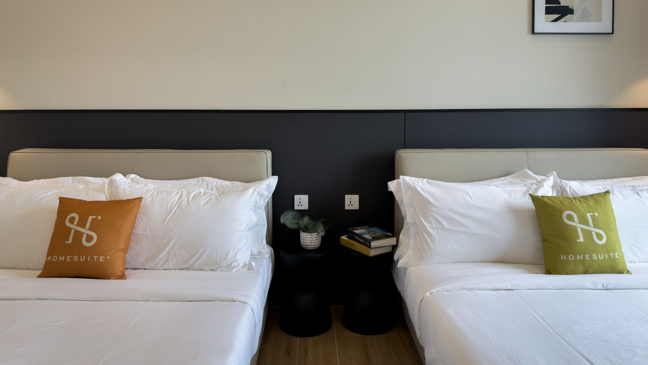 A hotel room with two beds, each with a pillow featuring the 'HOMESUITE' logo. There are bedside tables with books and a small plant.