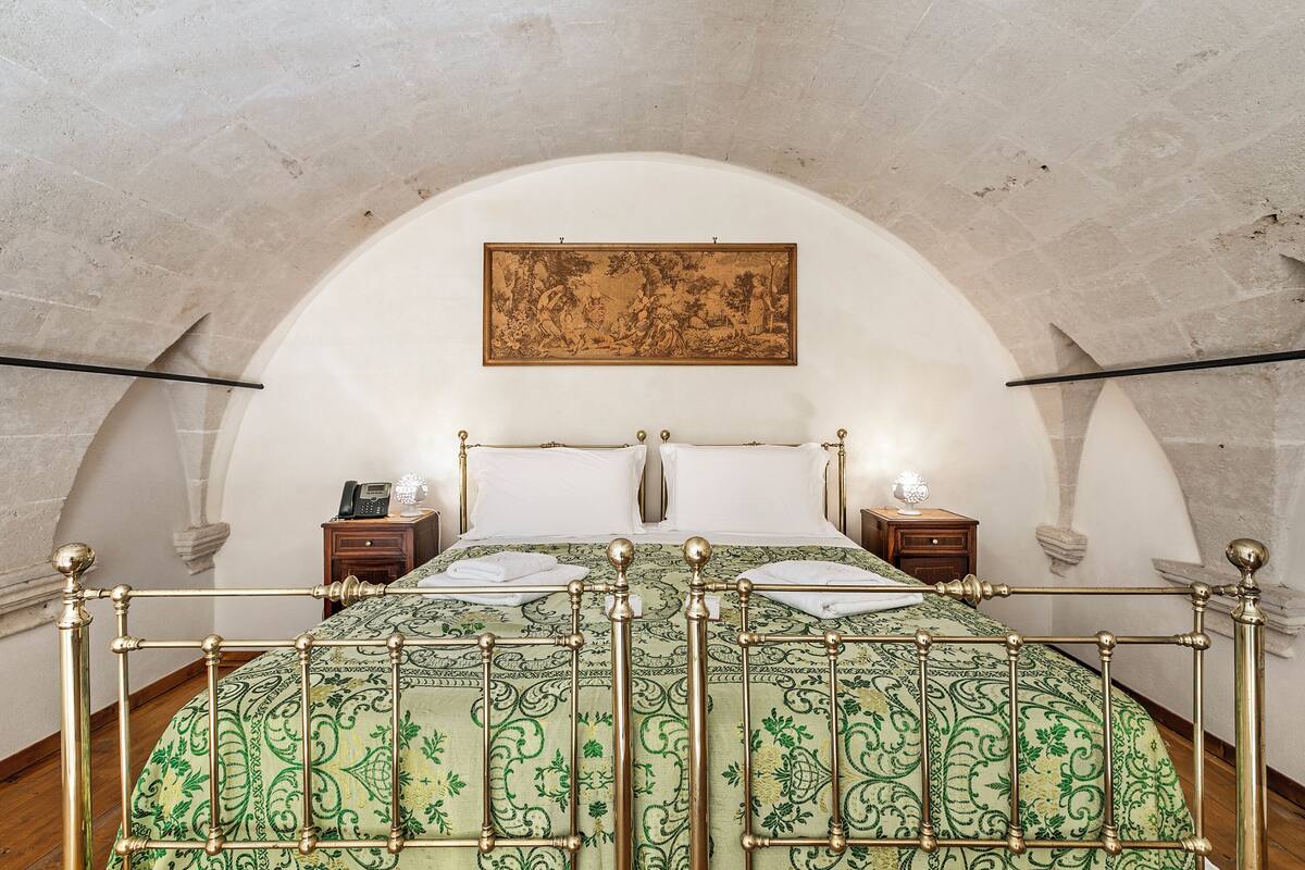 A bedroom with a high arched ceiling, a bed with a metal frame, two bedside tables with lamps, and a painting on the wall.