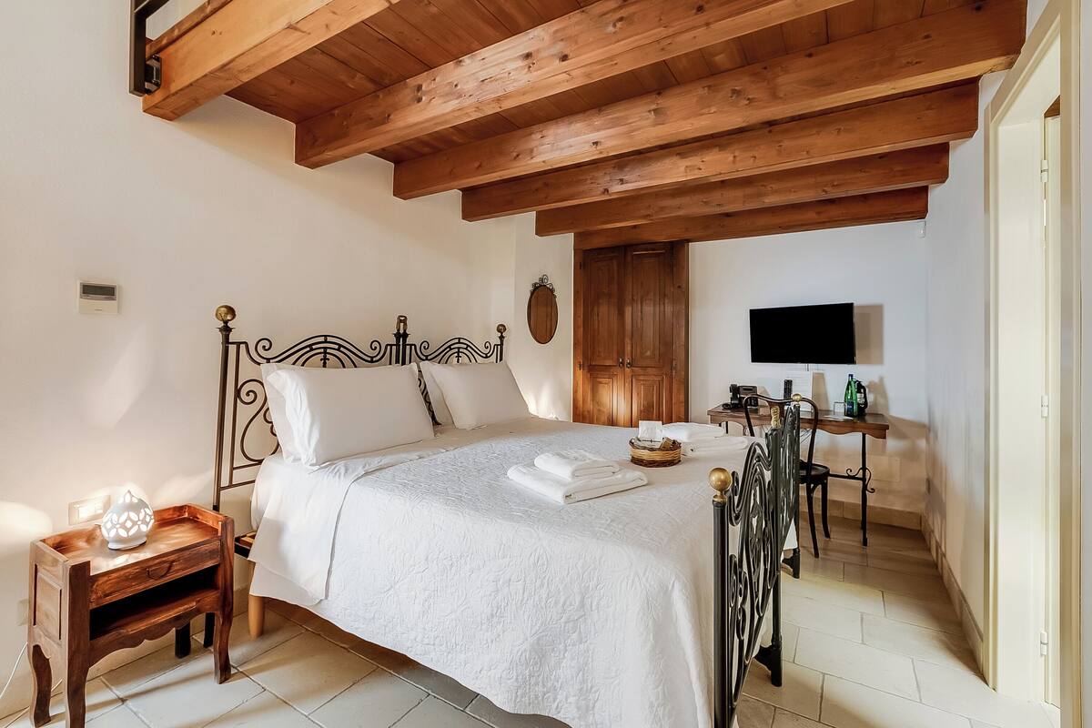 A bedroom with a wooden ceiling, a bed with white linens, a bedside table with a lamp, and a TV in the corner.