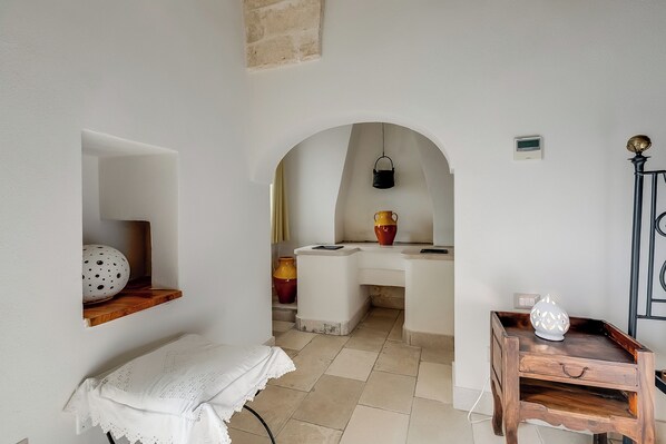 A room with a white wall, a wooden bench, a small table with a white vase, and a kitchen area with a sink and a hanging light. - Masseria Antoglia (Villa Castelli)