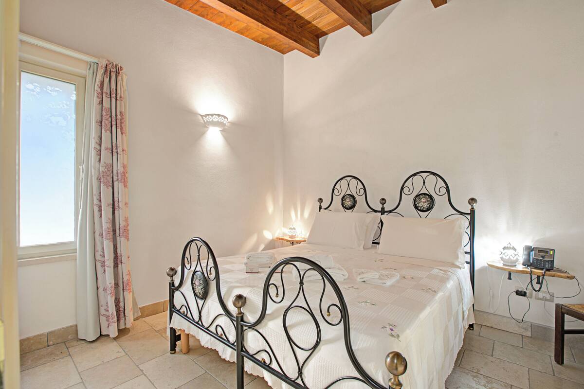 A bedroom with a bed, a bedside table, a nightstand, a window with curtains, and a wall-mounted light fixture.