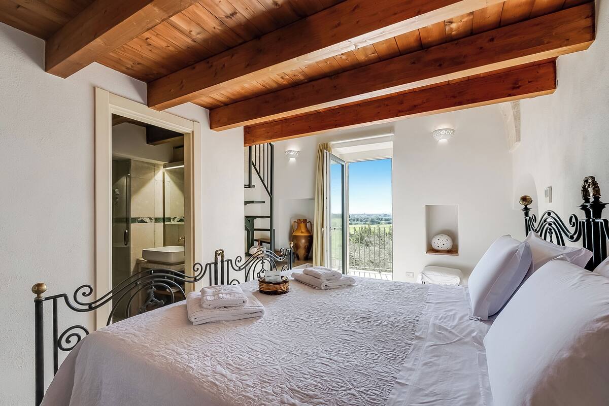 A bedroom with a wooden ceiling, a bed with white linens, a bathroom with a sink and mirror, and a view of the outdoors.